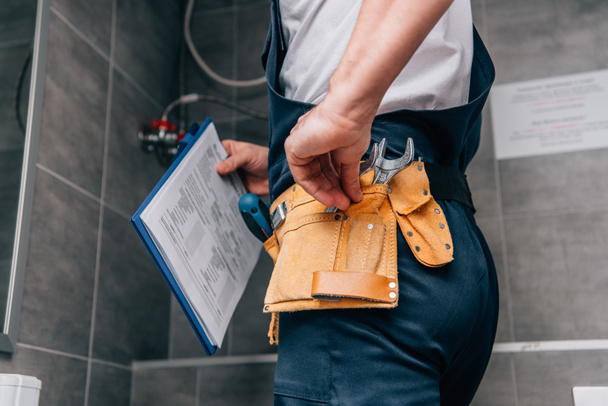 cropped shot of male plumber with clipboard and toolbelt standing in bathroom  - Φωτογραφία, εικόνα