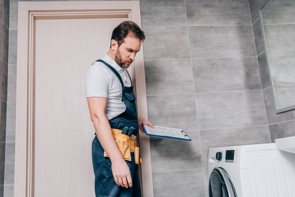 repairman with toolbelt and clipboard checking washing machine in bathroom - Photo, Image