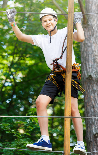 Niño en el parque de aventura forestal. El niño con casco naranja y camiseta azul sube por un sendero de cuerda alta. Habilidades de agilidad y escalada al aire libre centro de atracciones para niños. joven chico juega al aire libre
 - Foto, imagen