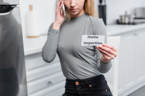 partial view of young woman holding card with lettering home inspection and talking on smartphone in kitchen  - Photo, Image