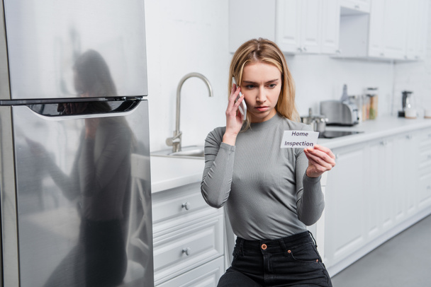 serious young woman holding card with lettering home inspection and talking on smartphone near broken refrigerator in kitchen  - Photo, Image