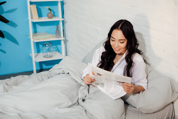szép, mosolygós, fiatal nő ült az ágyban, és az újság olvasás - Fotó, kép