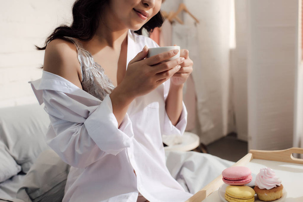 cropped shot of smiling asian girl in bra and white shirt holding cup of coffee and sitting on bed - Foto, immagini