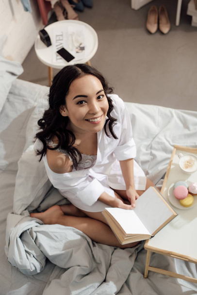 high angle view of beautiful happy asian girl sitting with book on bed and smiling at camera - 写真・画像