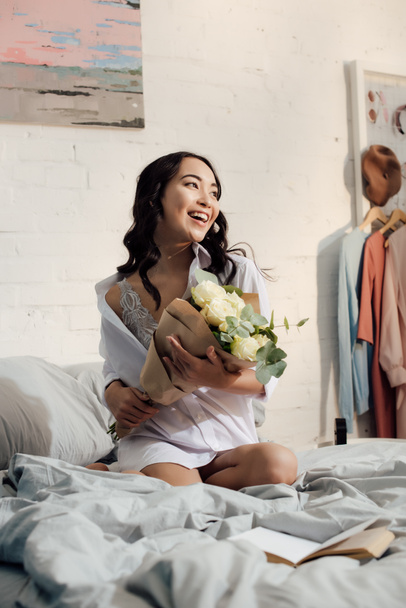 beautiful happy young asian woman in white shirt sitting on bed with bouquet of roses  - 写真・画像