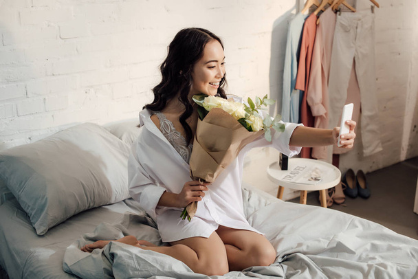 vue grand angle de belle fille asiatique heureuse tenant bouquet de fleurs et de prendre selfie avec smartphone dans la chambre
  - Photo, image