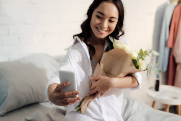 beautiful happy asian woman holding bouquet of roses and taking selfie with smartphone in bedroom - Φωτογραφία, εικόνα