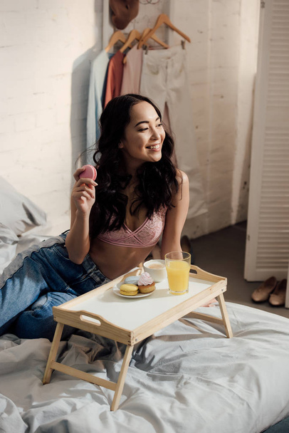 smiling young asian woman having breakfast and looking away in bedroom - Foto, Imagen