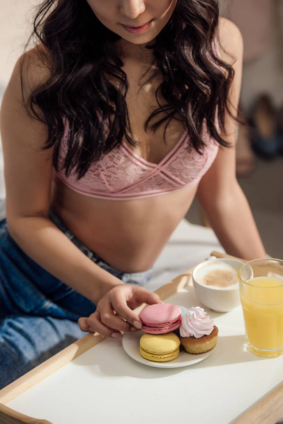 bijgesneden schot van sensuele jongedame in beha macarons voor ontbijt eten in slaapkamer - Foto, afbeelding