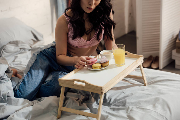 cropped shot of attractive girl holding tasty macaron while having breakfast in bedroom - Foto, Imagen