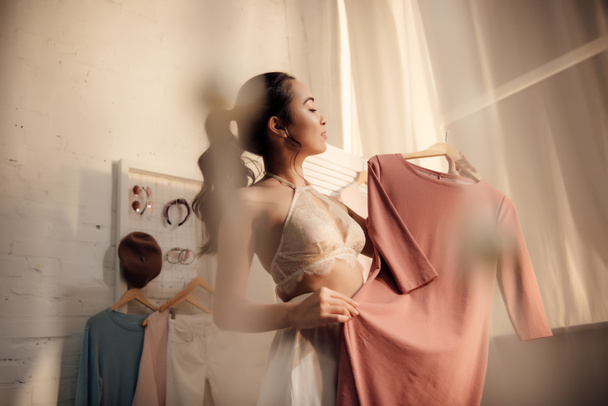 selective focus of beautiful asian woman in underwear holding hanger with stylish dress in wardrobe  - Foto, Imagen