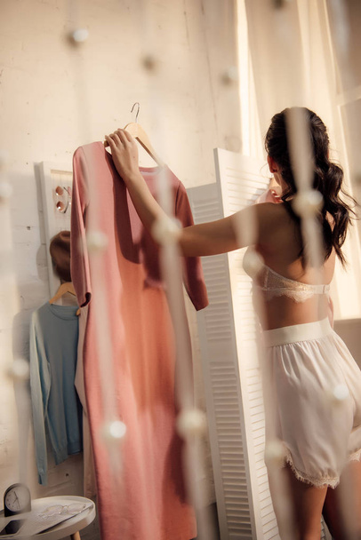 back view of young woman in underwear holding hanger with fashionable dress and looking away in wardrobe  - Foto, imagen