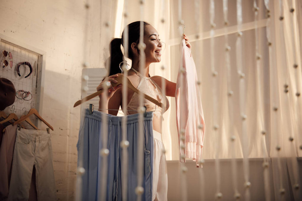 mooie jonge vrouw bedrijf hangers met stijlvolle kleren thuis glimlachen - Foto, afbeelding