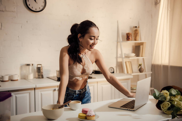 krásná usměvavá mladá žena lace bralette a džínách pomocí přenosného počítače během snídaně doma  - Fotografie, Obrázek