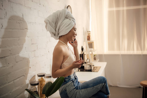 side view of attractive asian girl with towel on head talking by smartphone while sitting in kitchen - Photo, image