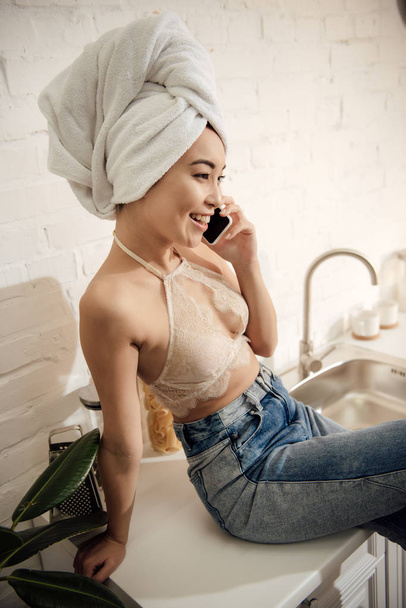 atractiva mujer asiática con toalla en la cabeza hablando por teléfono inteligente mientras está sentado en la cocina
 - Foto, imagen