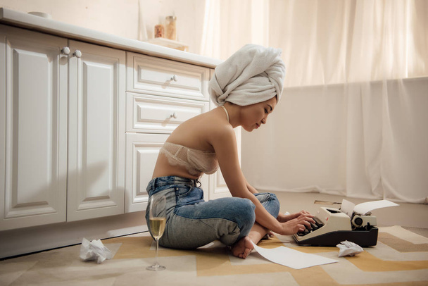 vista laterale di attraente donna asiatica con asciugamano sulla testa seduta nel pavimento e utilizzando macchina da scrivere
 - Foto, immagini