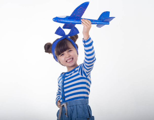 Beautiful child wearing striped dress holding blue airplane. Family vacation and tourism concept. White background, copy space - Фото, изображение