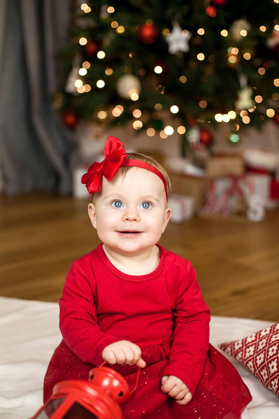 Little girl with blue eyes in a red dress sitting near the Christmas tree. Happy baby. Christmas homeliness. Holidays. - Foto, immagini
