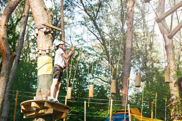 Child in forest adventure park. Kid in orange helmet  and blue t shirt climbs on high rope trail. Agility skills and climbing outdoor amusement center for children. young boy plays outdoors - Valokuva, kuva