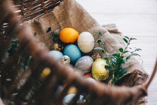 Stylish easter eggs on cloth in rustic basket on white wooden background. Easter hunt concept. Modern easter eggs painted with natural dye and green buxus branches. Happy Easter - Φωτογραφία, εικόνα