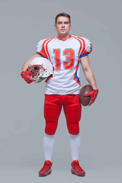 American football player holding rugby ball and helmet against grey background full length - Fotó, kép