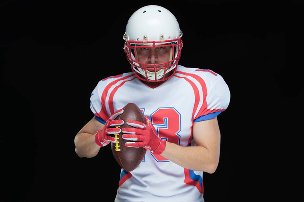 American football player wearing helmet posing with ball on black background - Foto, Imagen