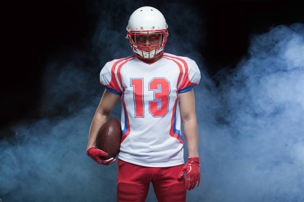 Front view portrait of american football player wearing helmet with ball against white smoke - Valokuva, kuva