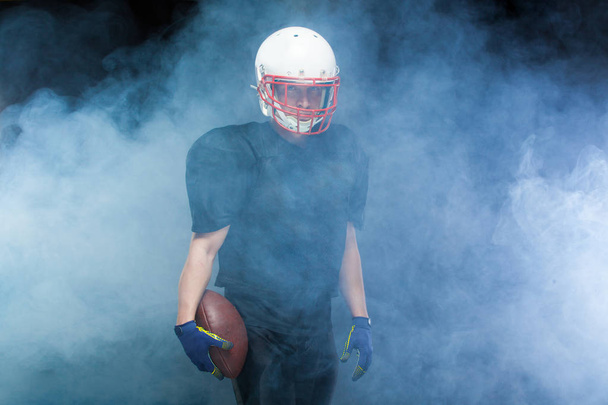 American football player in a black uniform, wearing helmet and holding ball against white smoke - Fotoğraf, Görsel
