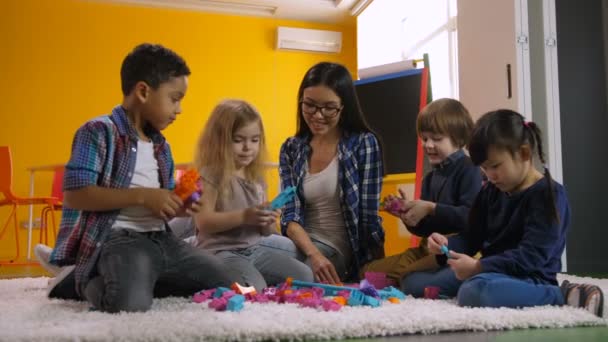 Kadehdittu monikansallinen monipuolinen lapset iloinen opettaja leikkii värikkäitä rakennuspalikoita lattialla päiväkodissa. Iloiset monikansalliset lapset, jotka nauttivat opetuspelistä esikoululuokassa
 - Materiaali, video