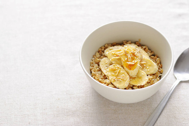 White ceramic bowl of oatmeal with banana slices and honey, served with the spoon on white table background. Viewed from high angle in closeup - Photo, Image