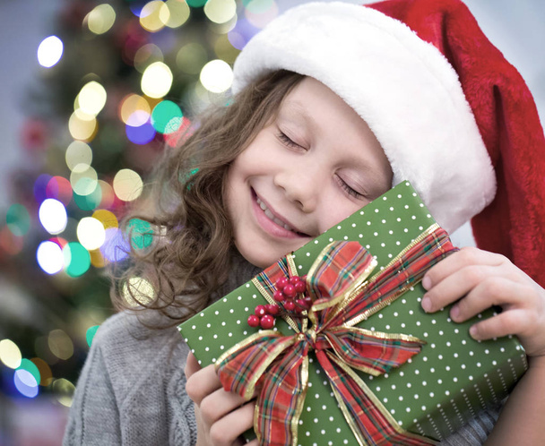 little girl in Santa hat holding a present under the Christmas tree - Фото, зображення