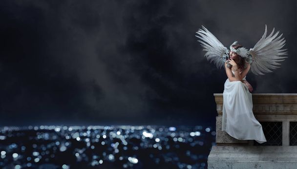 Νεαρή όμορφη γυναίκα στην Λευκή φόρεμα με φτερά αγγέλου κάθεται στην οροφή και να βλέπουν την πόλη το βράδυ. Όμορφη επαγγελματικό μακιγιάζ, τέλεια μαλλιά, κόμμωση με φυσικά φτερά και στρας. Χώρο αντίγραφο. - Φωτογραφία, εικόνα