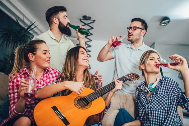Groep gelukkige jonge vrienden met gitaar met plezier en het drinken van bier in interieur  - Foto, afbeelding