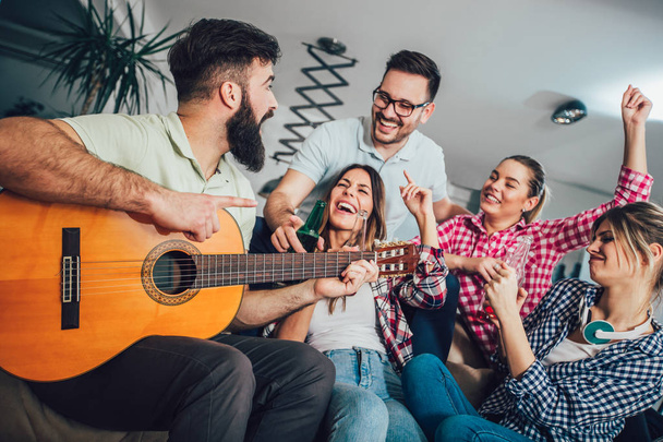 Group of happy young friends with guitar having fun and drinking beer in home interior  - Фото, изображение