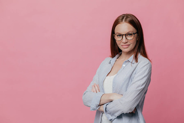 Sideways shot of prosperous businesswoman keeps arms folded, dressed in formalwear, wears spectacles, isolated over pink background with copy space for your promotion. Elegant teacher poses indoor - Photo, image