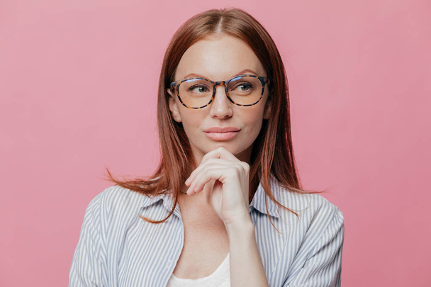 Contemplative attractive entrepreneur wears spectacles, formal shirt, keeps hand on chin, looks thoughtfully aside, thinks about new startup, poses against pink background. Close up horizontal shot - Photo, image