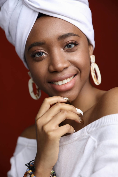 Beauty portrait of young smiling woman with blue contact lenses, dressed in white head tie, white dress and big earrings. Dark red background. - Photo, Image