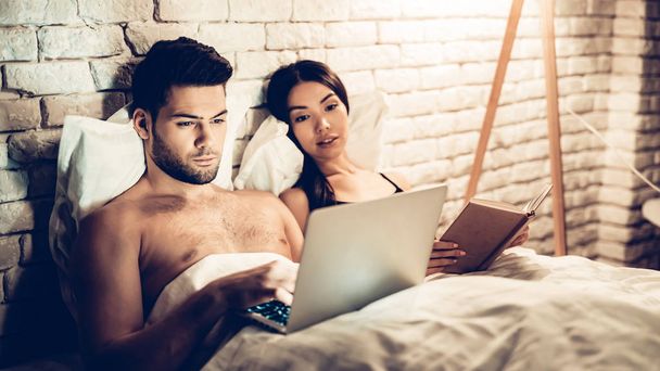 Couple Using Laptop Reading Book Before Sleeping - Φωτογραφία, εικόνα