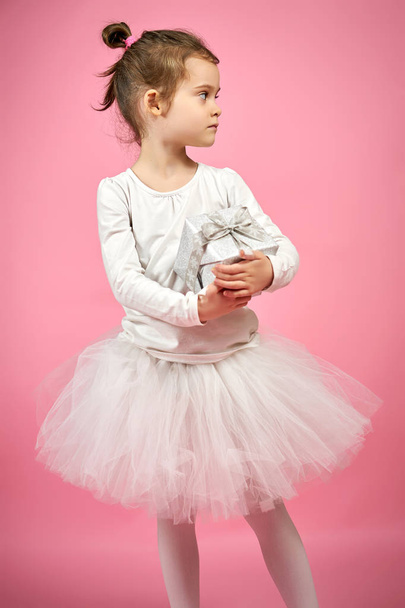 Милая маленькая девочка в юбке из тюля держит подарок на розовом фоне
 - Фото, изображение