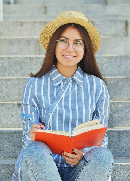 Portrait of a young Asian student in glasses with a hat dressed in a blue striped shirt that writes tasks and looks into the distance.Vertical photography - Фото, зображення