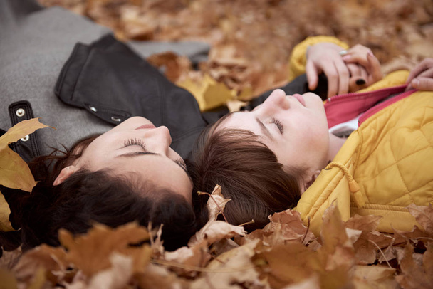 Pretty woman and teen girl are posing in autumn park. They are lying on fallen leaves. Beautiful landscape at fall season. - Foto, Bild