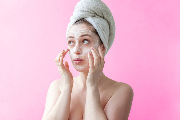 Beauty portrait of a smiling brunette woman in a towel on the head applying white nourishing mask or creme on face on pink background isolated. Skincare cleansing spa relax concept - Photo, Image