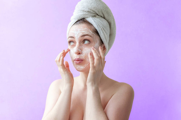 Portrait de beauté d'une femme brune souriante dans une serviette sur la tête en appliquant un masque nourrissant blanc ou crème sur le visage sur fond violet isolé. Soins de la peau nettoyage spa relax concept
 - Photo, image