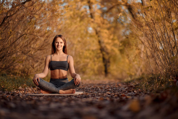Young woman practicing yoga exercise in autumn park with yellow leaves. Sports and recreation lifestyle. - Фото, изображение