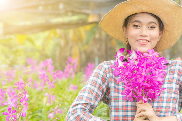 Kobieta ogrodnik w koszuli w kratę w kapeluszu trzyma różową orchideę w ręku. I uśmiechał się szczęśliwie. W ogrodzie orchidei - Zdjęcie, obraz
