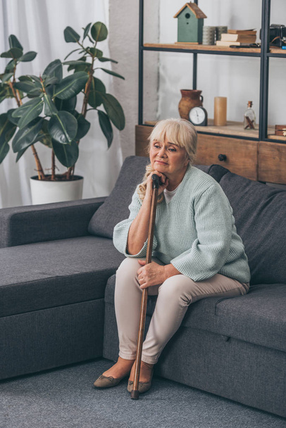 sad senior woman with blonde hair sitting with walking cane in living room  - Foto, Bild