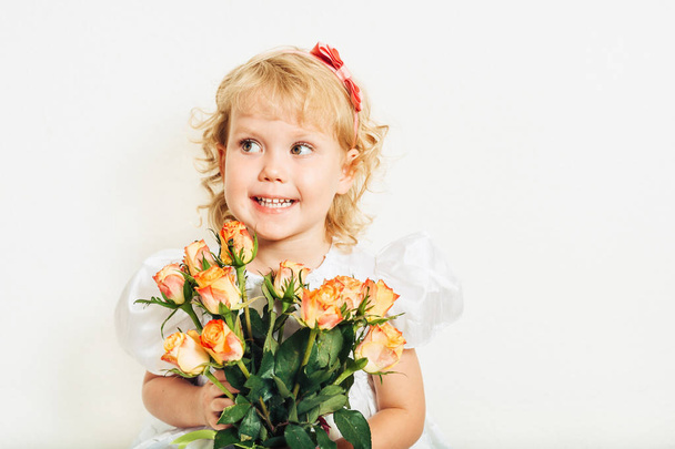 Studio shot of adorable 3 year old toddler girl wearing white occasion dress, holding bouquet of yellow orange roses - Photo, Image