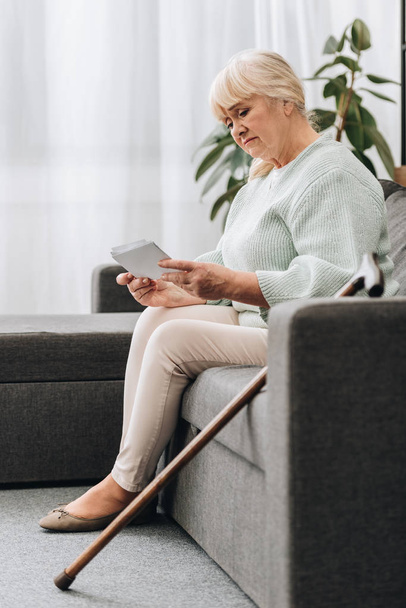 Traurige Rentnerin mit blonden Haaren schaut sich Fotos an, während sie auf dem Sofa sitzt - Foto, Bild