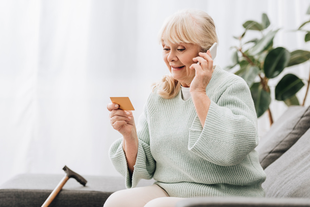 smiling blonde retired woman looking at credit card while talking on smartphone - Foto, Bild